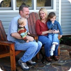 Couple sit outside on patio furniture. Each hold a child on their lap. 